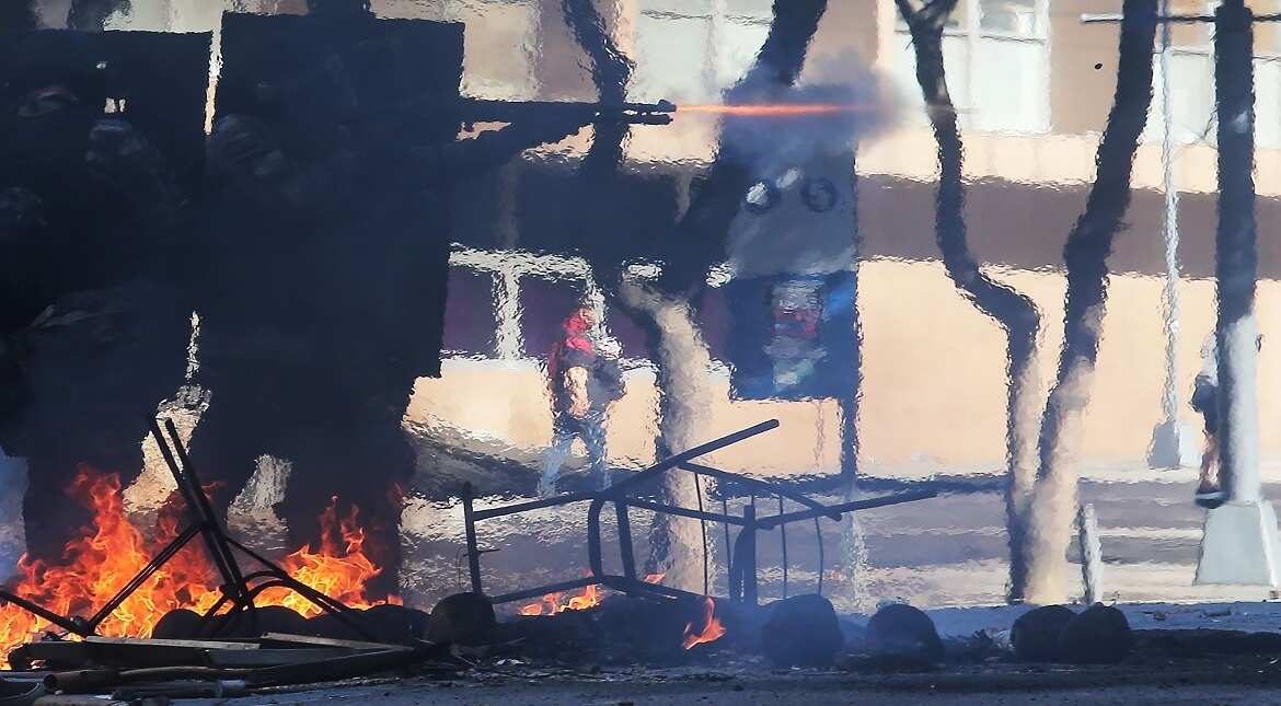 Military police fire a rubber bullet towards protesters as a street fire set by protesters burns